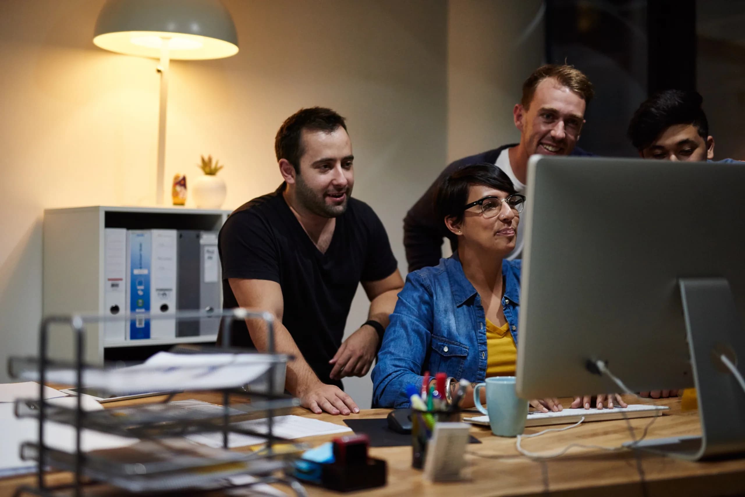 Four colleagues gathered around a desktop monitor in a dimly lit office, working through something on screen together