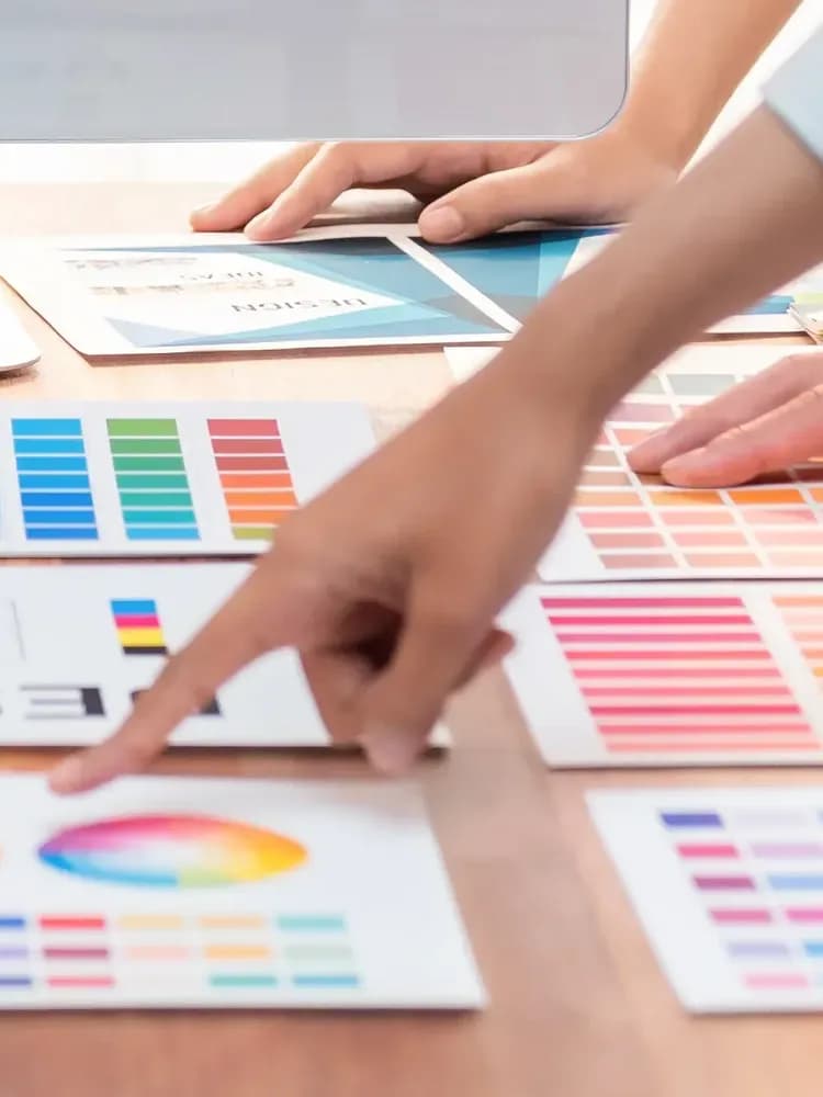 Hands pointing at printed colour swatches on a wooden table.